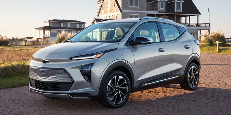 Silver 2024 Chevrolet Bolt EUV parked on a brick driveway in front of large houses with tall grass.