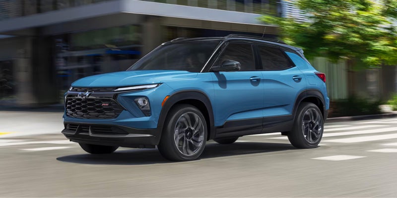 Blue Chevrolet Trax RS driving left across a crosswalk with blurred city buildings and trees in the background.